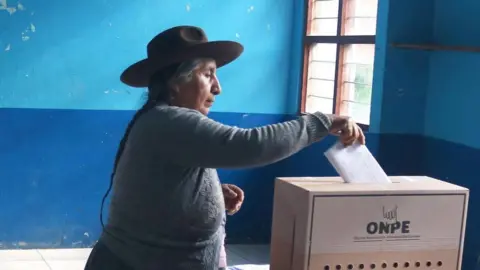 A woman with her hair in plaits and wearing a hat and a woolly jumper casts her vote during the presidential election in Urumbamba, Cusco, Peru, 12 April 2026. She is holding the ballot in her hand and is inserting it into a ballot box. 