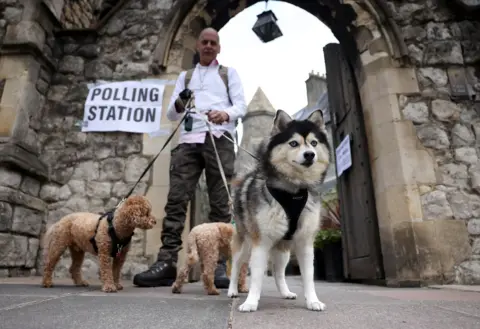 Neil Hall / EPA A large black and white dog with blue eyes, looking like a husky, stands looking directly at the camera with its ears perked, in front of two smaller brown, fluffy dogs, all on leads held by a person in a white shirt in front of a church door with a sign reading 'polling station'.