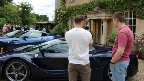 Wiltshire and Bath Air Ambulance Two men in jeans and t-shirts standing and looking at a black Ferrari with a large house, another ferrari and a large crowd and trees in the background. 