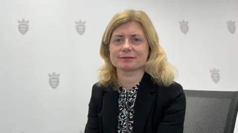 Deputy Lyndsay Feltham is sitting down and faintly smiling at the camera. She has shoulder-length blonde hair and blue eyes. She is wearing a black jacket over a black and white patterned blouse. Behind her the wallpaper features the Jersey shield of arms design.