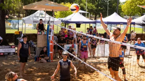 Kaktus Scot A man jumping in the air with both arms raised to hit a ball over a beach volleyball net with players and crowds watching