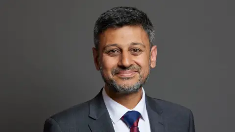 PA Dr Zubir Ahmed smiles, looking into the camera. He is wearing a white shirt, grey blazer and a blue and red tie