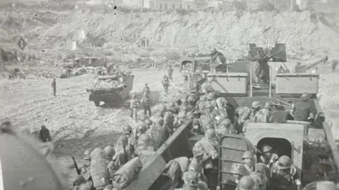 Victor Todd A black and white photo of American troops making their way off a large landing craft wearing full military kit. The photo was taken by the ships commander from the quarter deck.