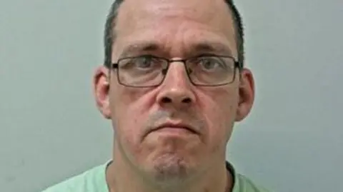 Police handout Custody image of Gary Wanless wearing black glasses and bright green t-shirt.