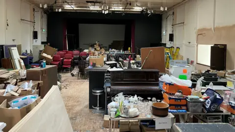 A large hall is full of clutter - including chairs, card boxes, glasses, crates, tables a piano. A dark stage curtain can be seen at the far end of the hall. The parquet flooring is badly in need of repair. 