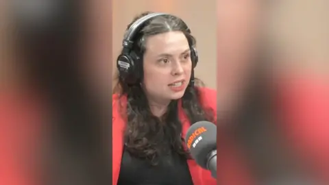 A woman with dark, curly hair talks to someone off-camera.