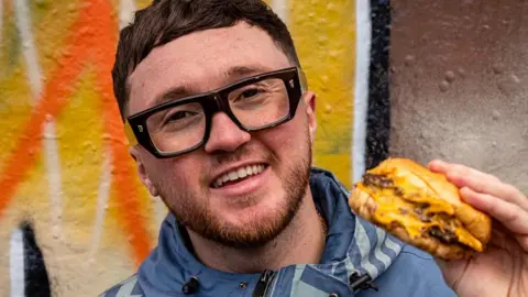 Oscar is wearing dark rimed glasses and is smiling at the camera whilst holding up a cheeseburger. He's standing in front of orange graffiti.