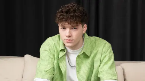 Sam Meyer, who is wearing a white T-shirt with a lime green shirt and a silver chain, is staring directly at the camera while sitting on a cream couch. 