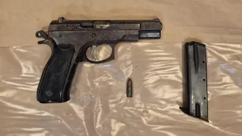An Garda Síochána A black handgun and one bullet lying on top of an evidence bag as part of a display. The bag is made of brown paper and clear plastic.