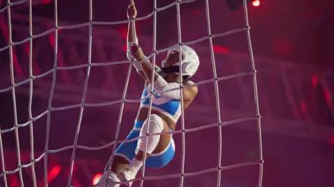 BBC/Hungry Bear Media Emily Bell in a blue and white sports kit and white safety helmet climbs a cargo net during the eliminator event.