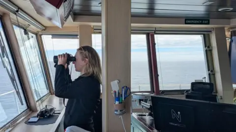 Orca Ellie Adams, a young woman with shoulder-length blonde hair wearing a black top , is on the bridge of a boat looking out to sea.