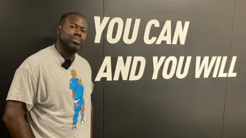 James Grant/BBC Swifty wearing a T-shirt in front of a sign that read "you can and you will"