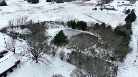 A drone image of a pond, with a thin layer of ice over part of it, near some houses