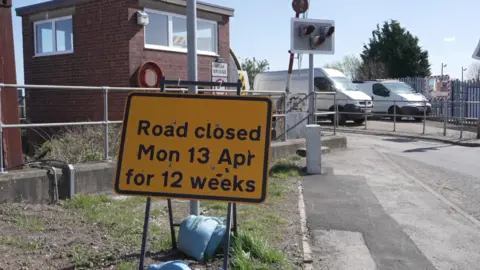 A yellow road sign reading "Road closed Mon 13 Apr for 12 weeks". A red brick building and two vans can be seen in the background. 