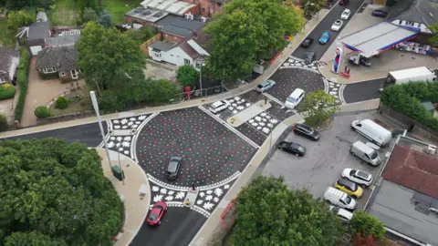 BBC An aerial photo of two roundabouts, with rainbow leaves painted on the tarmac. At each of the the three exists for each roundabout there's a band of white leaves painted across the road.
