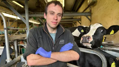 Daniel Chivers A farmer with short brown hair and wearing a quarter-zip hoodie with his arms crossed and wearing blue plastic gloves, standing in a milking parlour with dairy cows and milking equipment in the background.
