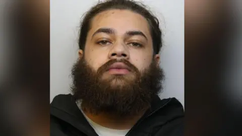 A mugshot of Jardel Edwards - he is wearing a white T-shirt and black hoodie and has a beard and brown hair.