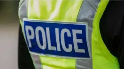 The back of a police officer's hi-vis jacket. It it florescent yellow and has the word police in white capital letters on a blue background on it. 