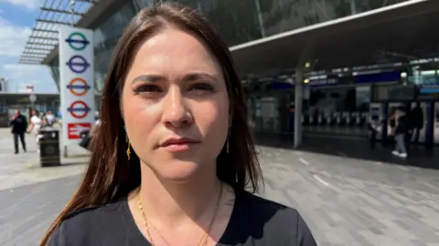 Esme Rice stands on the station concourse at Stratford. She is a young woman with long, dark hair and wears a black top.