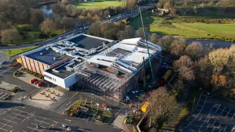 Aerial view of the Sands Centre taken in January after the storm. The right-hand side of the venue is covered with scaffolding, as is the section of roof above. Parts of a temporary canopy appear to have blown off. To the front and side of the building is a car park, while fields, a river and trees can be seen in the background.