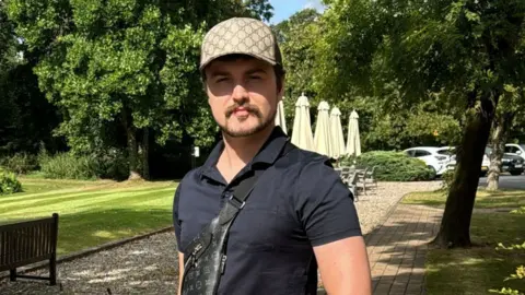 Osian is standing in the middle of a gravel path with grass either side of him. Behind him are tables with umbrellas folded up. Osian is wearing a beige baseball cap and a navy polo shirt, with a black bag across his chest with a Louis Vuitton symbol pattern over it. He has a brown goatee with a short beard.