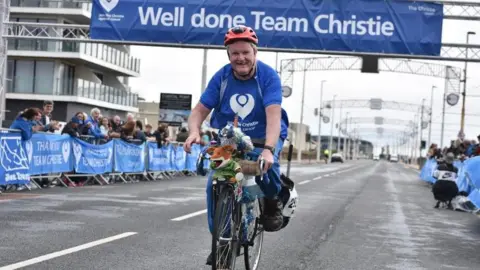 Tony O'Dwyer, wearing a blue Christie T-shirt, cycles along Blackpool Promenade with a Basil Brush mascot on the front of his bicycle.