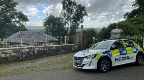 Un automóvil policial estacionado al costado de una carretera. Detrás hay una pared con barandas blancas con púas. Detrás de la pared se pueden ver el techo y las chimeneas de un edificio