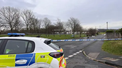 A police car is in the foreground on a road. Police tape is in the background and the cordon includes an area of grass, pavements and a road.  