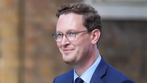 Darren Jones, a young man with glasses, wearing a blue suit and tie, smiles as he walks up Downing Street