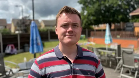 Ben Schofield/BBC George Jeffery looking directly to the camera. He is standing outside on a bright day, with the sun shining on the left side of his face. Behind him are tables and chairs. He is wearing a striped, collared oolo shirt and has light brown hair, cut relatively short.