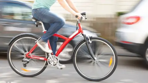 Action shot of a man riding a red bike, he is wearing denim jeans and green tshirt, there are two silver cars blurred in the background