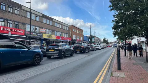 Traffic queues along the left-hand lane of a busy high street in Bury Park, Luton, with pedestrians and trees on the pavement to the right.