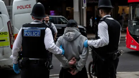 Reuters Two police officers in uniform with their backs to the camera hold a handcuffed suspect by the arms as they guide him towards a police van. 