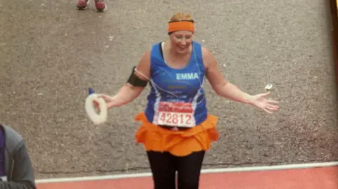 Emma Smith A woman dressed in a blue running top, black leggings and orange headband crosses a finish line holding a circular water bottle