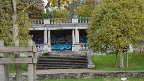 Shaun Whitmore/BBC A grey stone pavilion, viewed from a distance with a tree in the left foreground. The pavilion is above a flight of terraced stone steps. It is fronted by grey columns. Across its roof is a row of smaller, tenpin-shaped columns topped by a stone rail, creating a balcony. The back wall of the pavilion is covered in graffiti. 