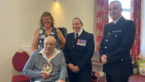 Eric Birkin, in a wheelchair, holding a police plaque. A woman in a floral dress is standing behind him, with two police officers, Ch Supt Claire Talbot and Det Ch Insp Rob Huddleston, both in police uniform, standing by him and smiling.