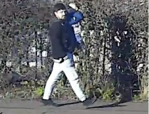 Police Scotland A man with dark hair wearing a dark jacket and white trousers carries a young child dressed in dark blue clothing. They appear to be walking along a pavement with a bush seen in the background.