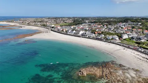 Cobo bay in Guernsey viewed from above. You can see the white sand and turquoise water. There are cars travelling along the road which is behind the beach and you can see houses and fields stretching off into the distance. 