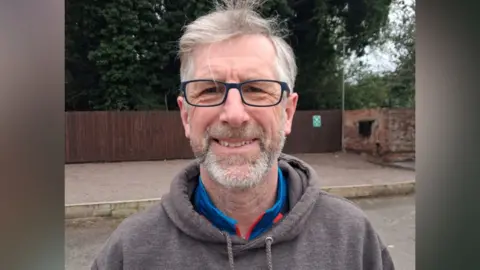 Supplied A bespectacled man with grey hair and beard stood outside in a brown hoody