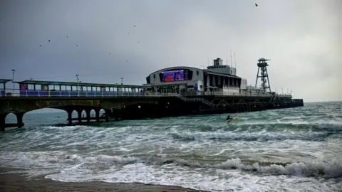 Moxley A swimmer with a surfboard in the choppy sea next to a pier.