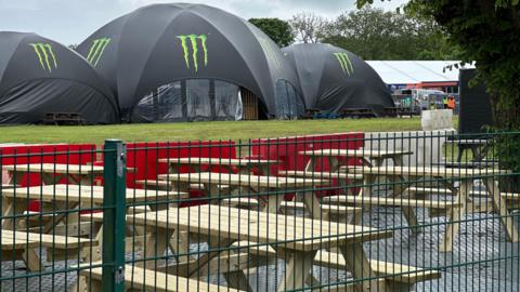Picnic tables behind a fence with three large Monster branded tents behind