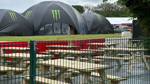 BBC Picnic tables behind a fence with three large Monster branded tents behind