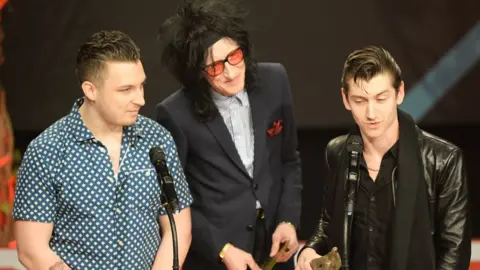 Getty Images Arctic Monkeys Drummer Matt Holders dan John Cooper Clarke berdiri di sebelah vokalis Arctic Monkeys Alex Turner saat ia menyampaikan pidato di NME Awards 2014