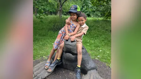 Contributor Picture Two children are sitting on a wooden sculpture of a mole. The girl is wearing a baseball hat and a floral summer dress. The boy is in a beige polo shirt and matching shorts. 