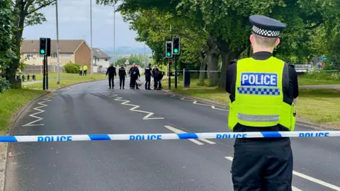 A police man stands with his back to the camera. He is standing behind blue and white police tape. In the distance a group of officers in black uniforms clear a scene on an empty road