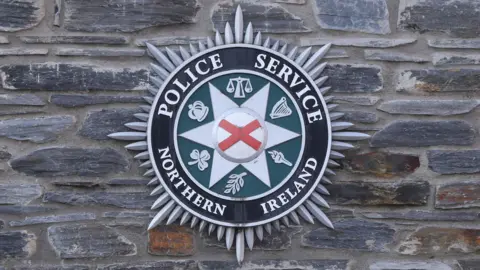 Black, green, and grey circular crest on a grey brick wall, featuring the words Police Service Northern Ireland in white lettering.