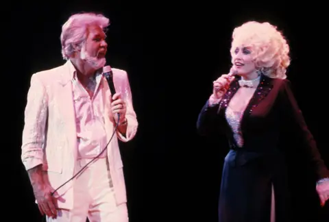 PL Gould/IMAGES/Getty Images Kenny Rogers, in a white suit, and and Dolly Parton in a dark fitted suit top over a white bustier, sing together 