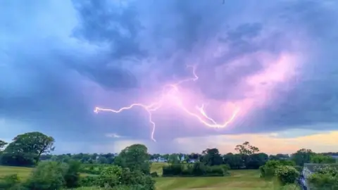 Chris Cumulus/BBC Weather Watchers Strikes of lightning in St Andrew Guernsey. The lightning is purple coloured on a blue/grey coloured skyline. There is also greenery and trees visible.