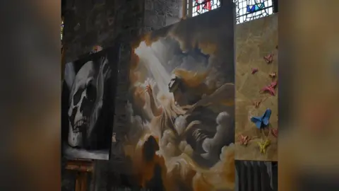 Three paintings depicting different artworks are displayed inside St Cuthbert's Church. There are stained windows in the background.