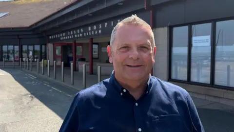 A middle-aged man grinning at the camera. He is standing outside a hospital. The man is wearing a smart black shirt. It's a sunny day.